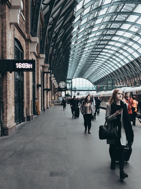 Inside a busy train station with a large curved glass and steel roof arching overhead, several pedestrians walk along the platform. In the foreground, a young woman with long blonde hair, dressed in a